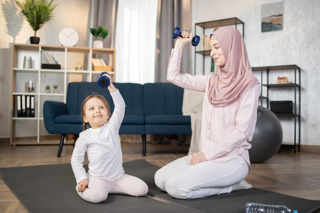 Jeune femme arabe heureuse faisant de l'exercice le matin avec sa belle petite fille à la maison assise sur un tapis