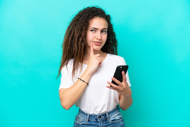 Jeune femme arabe isolée sur fond bleu à l'aide d'un téléphone portable et de la pensée