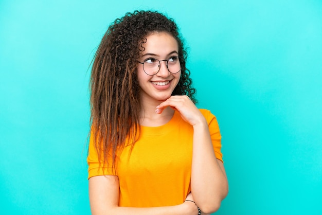 Jeune femme arabe isolée sur fond bleu avec des lunettes et pensant en levant les yeux