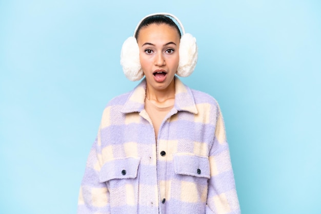 Jeune femme arabe portant des manchons d'hiver isolé sur fond bleu avec une expression faciale surprise