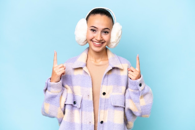 Jeune femme arabe portant des manchons d'hiver isolés sur fond bleu pointant vers le haut une excellente idée