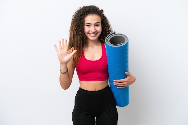 Jeune femme arabe sportive allant à des cours de yoga tout en tenant un tapis isolé sur fond blanc en comptant cinq avec les doigts