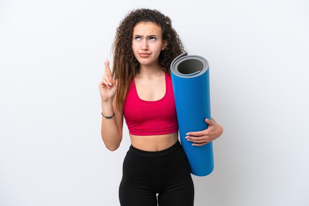 Jeune femme arabe sportive allant à des cours de yoga tout en tenant un tapis isolé sur fond blanc avec les doigts croisés et souhaitant le meilleur