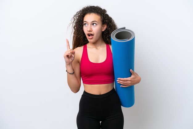 Jeune femme arabe sportive allant à des cours de yoga tout en tenant un tapis isolé sur fond blanc en pensant à une idée pointant le doigt vers le haut