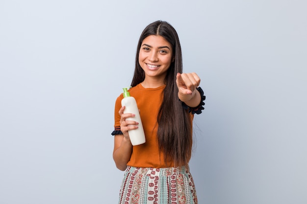 Jeune femme arabe tenant une bouteille de crème sourires joyeux pointant vers l'avant.