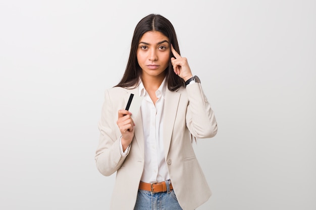 Jeune femme arabe tenant une carte de crédit pointant sa tempe avec le doigt, pensant, concentrée sur une tâche.