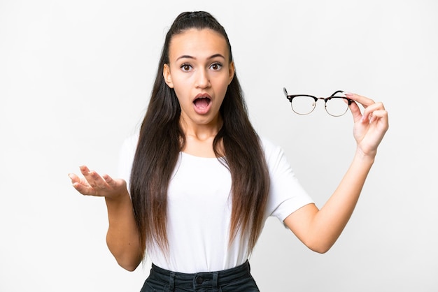 Jeune femme arabe tenant des lunettes sur fond blanc isolé avec une expression faciale choquée