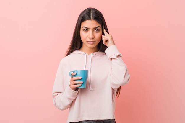 Jeune femme arabe tenant une tasse pointant sa tempe avec le doigt, pensant, concentrée sur une tâche.