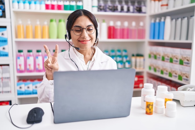 Jeune femme arabe travaillant à la pharmacie à l'aide d'un ordinateur portable montrant et pointant vers le haut avec les doigts numéro deux tout en souriant confiant et heureux.