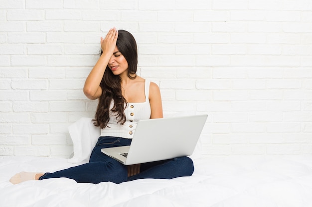 Jeune femme arabe travaillant avec son ordinateur portable sur le lit en oubliant quelque chose, en frappant le front avec la paume et en fermant les yeux.