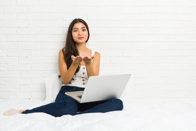 Jeune femme arabe travaillant avec son ordinateur portable sur le lit tenant quelque chose avec des paumes, offrant à la caméra.