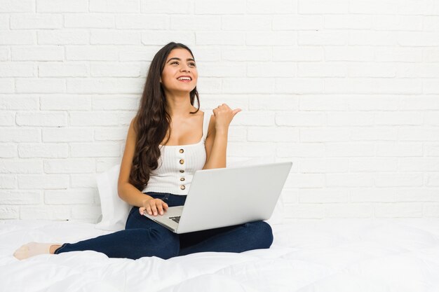 Jeune femme arabe travaillant avec son ordinateur portable sur les points du lit avec le doigt de pouce loin, riant et insouciant.