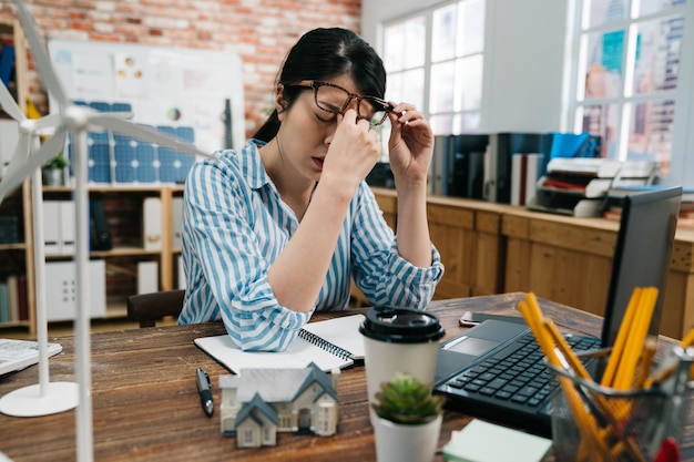 Jeune femme architecte asiatique élégante dans des verres fatigués se frottant le nez et les yeux se sentant fatigués et mal à la tête. Concept de stress et de frustration surmené sur le plan de construction du bâtiment de l'économie verte.