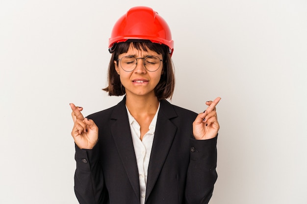 Jeune femme architecte avec casque rouge isolé sur fond blanc croisant les doigts pour avoir de la chance