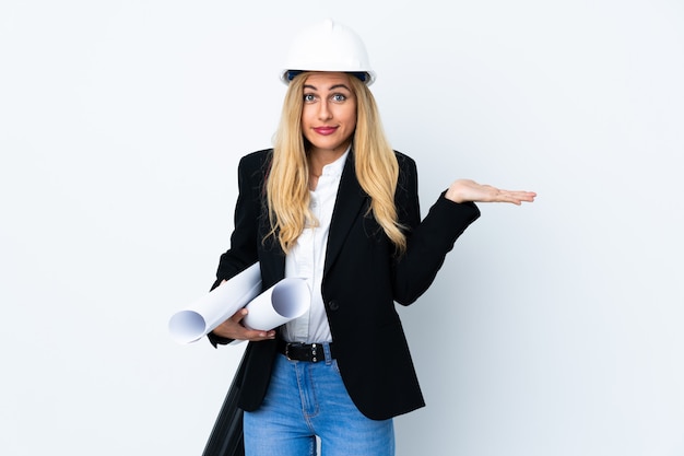 Jeune femme architecte avec casque et tenant des plans sur blanc isolé ayant des doutes avec l'expression du visage confus