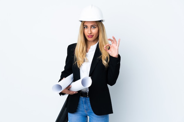 Jeune femme architecte avec casque et tenant des plans sur blanc isolé montrant un signe ok avec les doigts