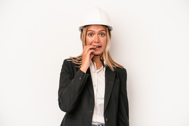 Jeune femme architecte caucasienne portant un casque isolé sur fond blanc se rongeant les ongles, nerveuse et très anxieuse.