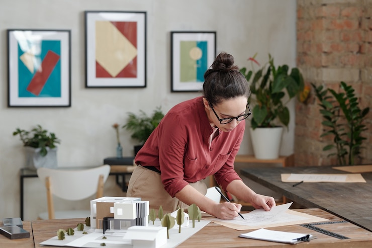 Photo jeune femme architecte occupée avec un chignon debout à table avec un modèle 3d de maison de campagne et éditant le plan de construction
