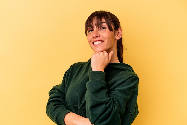 Jeune femme argentine isolée sur fond jaune souriante heureuse et confiante, touchant le menton avec la main.