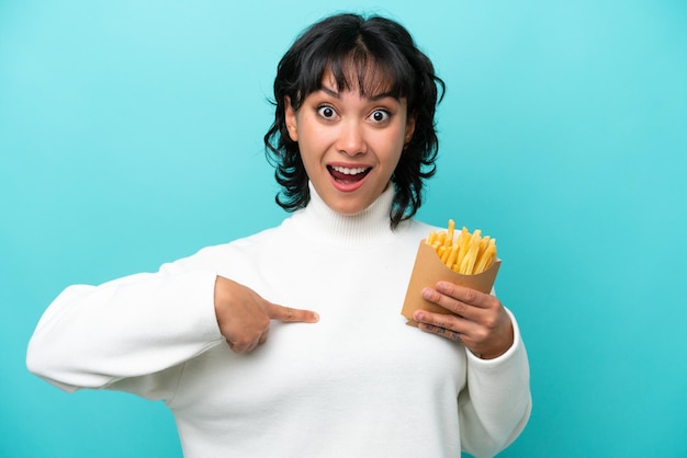 Jeune femme argentine tenant des frites frites isolées sur fond bleu avec une expression faciale surprise