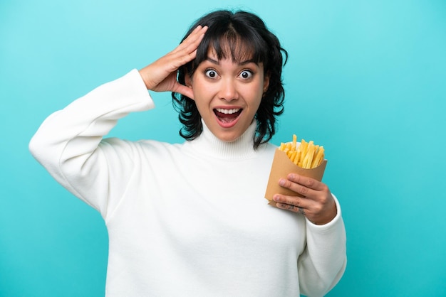 Jeune femme argentine tenant des frites frites isolées sur fond bleu avec une expression de surprise