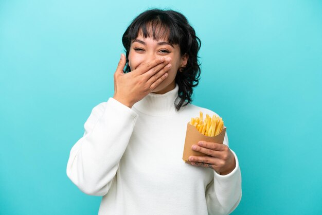 Jeune femme argentine tenant des frites frites isolées sur fond bleu heureuse et souriante couvrant la bouche avec la main