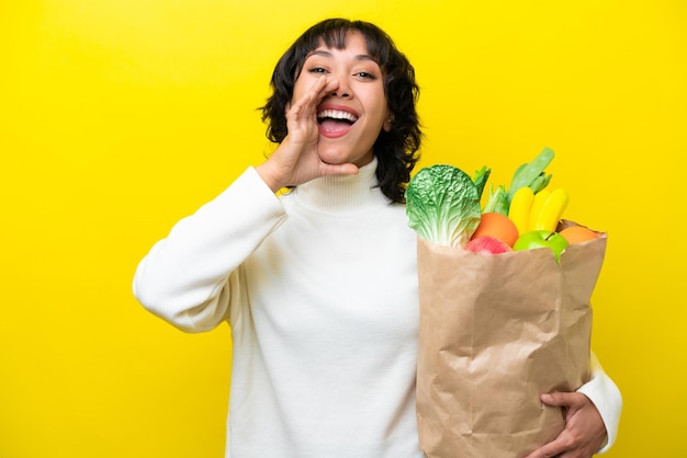 Jeune femme argentine tenant un sac d'épicerie isolé sur fond jaune criant avec la bouche grande ouverte