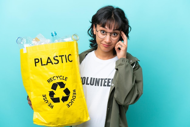 Jeune femme argentine tenant un sac plein de bouteilles en plastique à recycler isolé sur fond bleu en pensant à une idée