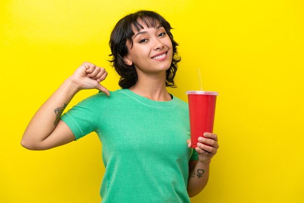 Jeune femme argentine tenant un soda isolé sur fond jaune fière et satisfaite