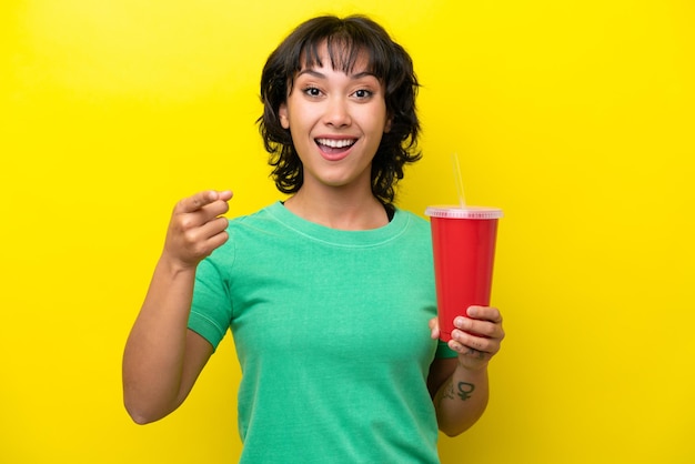 Jeune femme argentine tenant un soda isolé sur fond jaune surpris et pointant vers l'avant