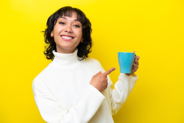 Jeune femme argentine tenant une tasse de café isolée sur fond jaune et la pointant