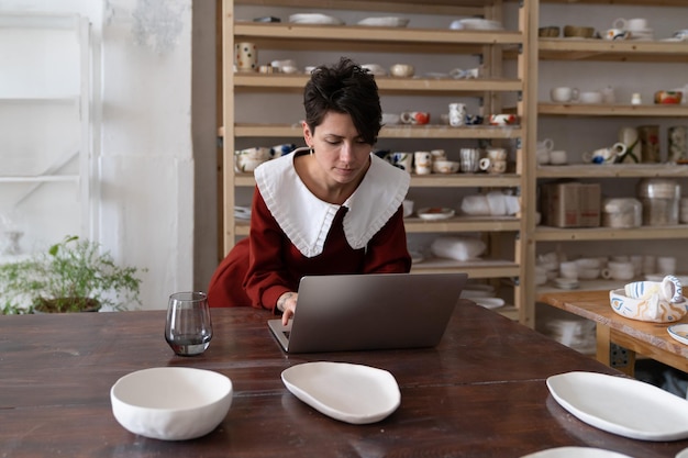Jeune femme artisane travaillant sur un ordinateur portable dans un studio de poterie gagner de l'argent avec de la céramique