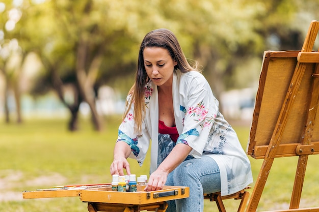 Jeune femme artiste arrangeant des pots de peinture sur une table alors qu'elle était assise devant une toile dans le parc.