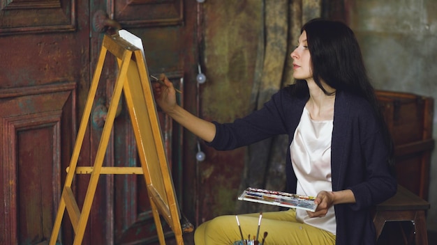 Photo jeune femme artiste dessiner une image avec des peintures à l'aquarelle et un pinceau sur une toile de chevalet