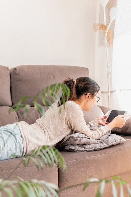 Jeune femme asiatique allongée sur le canapé à la maison, regarder des vidéos et des films sur une tablette