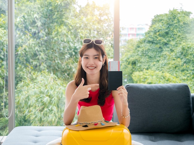 Jeune femme asiatique assez heureuse avec des lunettes de soleil montre un passeport et un billet d'avion avec un sourire assis sur un canapé près d'une valise jaune de chapeau de plage et d'une fenêtre en verre prête à voyager sur le concept de vacances d'été