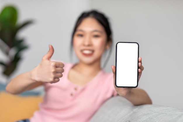 Jeune femme asiatique assise sur un canapé à la maison montrant un smartphone avec un écran blanc et un pouce vers le haut