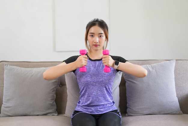 Une jeune femme asiatique assise sur le canapé pour faire de l'exercice avec des haltères pour entraîner des bras forts