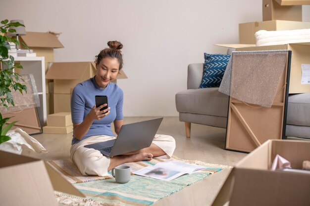 Photo une jeune femme asiatique assise dans une nouvelle maison en utilisant un smartphone et un ordinateur portable achète des décorations en ligne après avoir emménagé