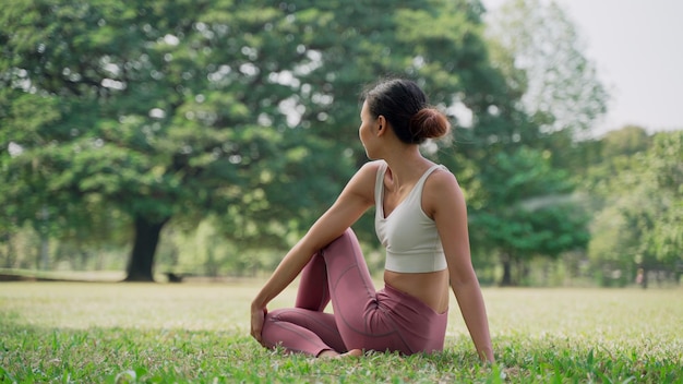 Jeune femme asiatique assise sur l'herbe dans la DEMI-SEIGNEURS DES POISSONS POSE à l'extérieur dans le parc de la ville avec le fond des grands arbres. Vue arrière d'une femme pratiquant le yoga à l'extérieur par une journée ensoleillée.