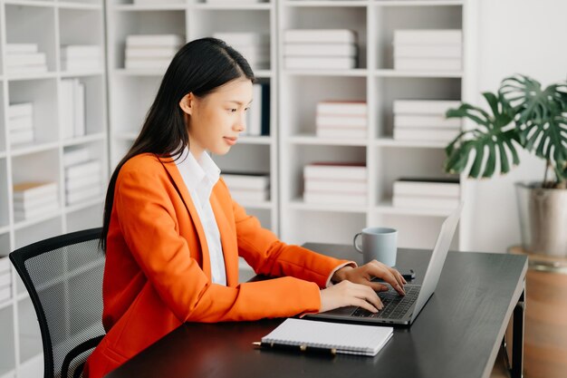Jeune femme asiatique attrayante, employée de bureau, costumes d'affaires, souriant à la caméra avec un bloc-notes, une tablette et un ordinateur portable au bureau.