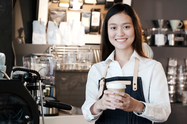 Jeune femme asiatique Barista tenant une tasse de café à emporter avec un visage souriant au café comptoir