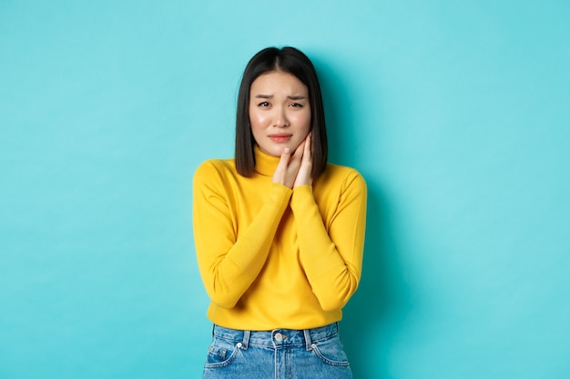 Jeune femme asiatique a besoin d'un dentiste, touchant la joue et fronçant les sourcils de maux de dents douloureux, debout sur fond bleu. Concept de soins de santé.