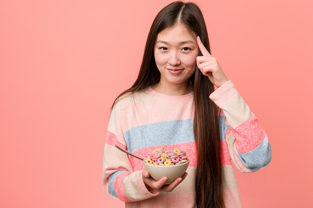 Jeune femme asiatique avec un bol de céréales pointant sa tempe avec le doigt, pensant, concentrée sur une tâche.