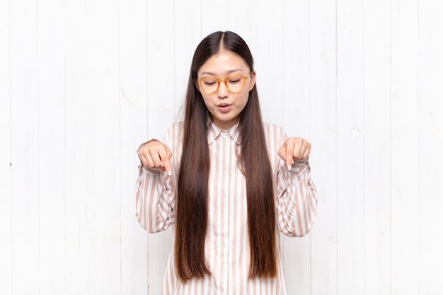 Jeune femme asiatique avec la bouche ouverte pointant vers le bas avec les deux mains, l'air choqué, étonné et surpris