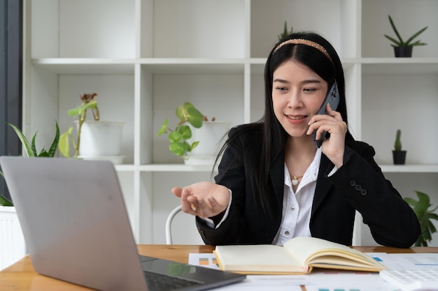 Jeune femme asiatique de bureau parlant à quelqu'un sur son téléphone portable tout en regardant au loin avec une expression faciale heureuse