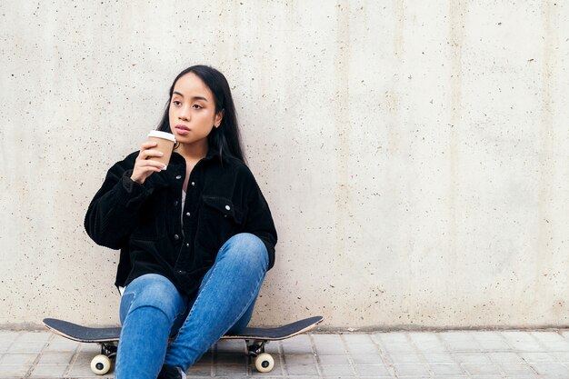 Jeune femme asiatique buvant du café à l'extérieur assise sur sa planche à roulettes appuyée contre un mur de béton