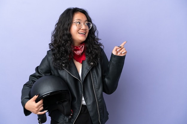 Jeune femme asiatique avec un casque de moto isolé sur fond violet dans l'intention de réaliser la solution tout en levant un doigt vers le haut