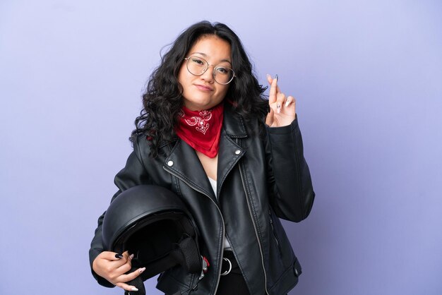 Jeune femme asiatique avec un casque de moto isolé sur fond violet avec les doigts croisés et souhaitant le meilleur