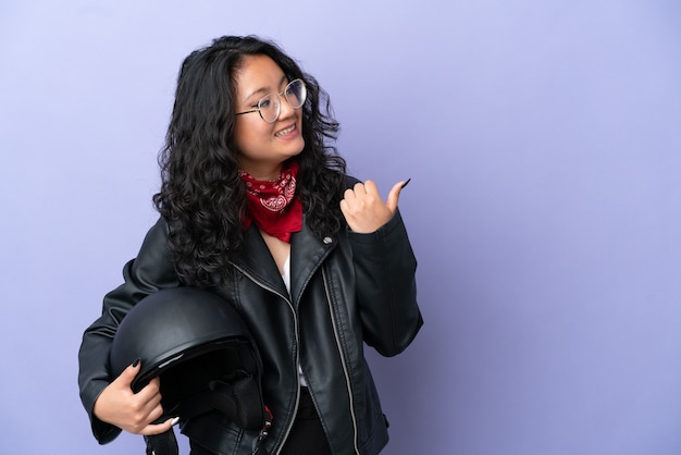 Jeune femme asiatique avec un casque de moto isolé sur fond violet pointant vers le côté pour présenter un produit